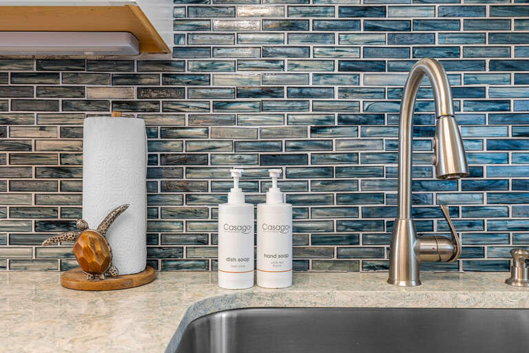 Enjoy the fresh, beachy vibes in this well-equipped coastal kitchen featuring ocean-inspired tile, stainless fixtures, and complimentary Casago essentials for your perfect seaside stay.