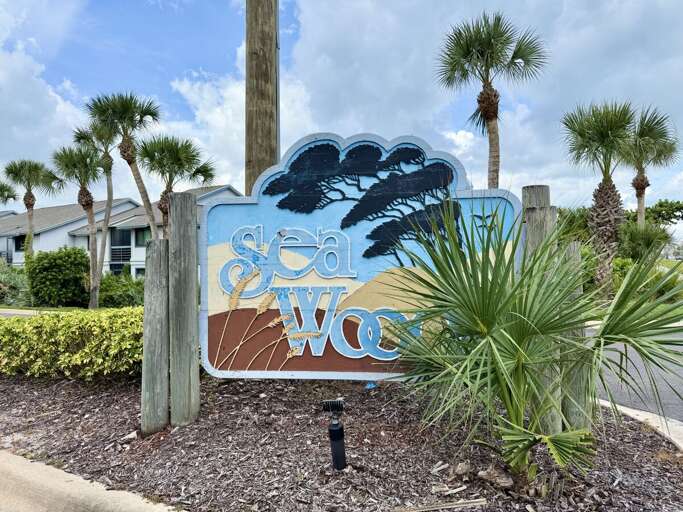 Entrance sign surrounded by palm trees and coastal landscaping at Sea Woods. This inviting beach stay community offers a relaxed seaside atmosphere just moments from sandy shores and ocean breezes.
