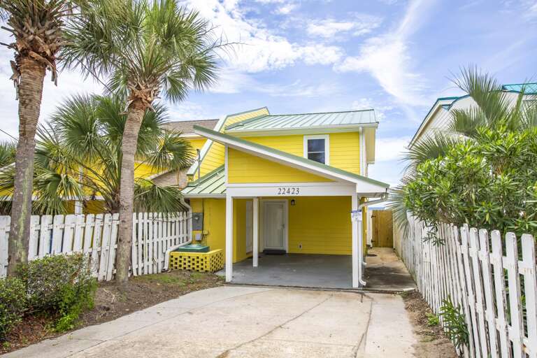 Yellow two-story house with green metal roof, covered carport, white picket fence, and palm trees.