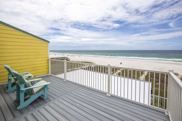Expansive gray composite deck with aqua Adirondack chairs and striped cushions beside a yellow exterior wall overlooking the ocean.