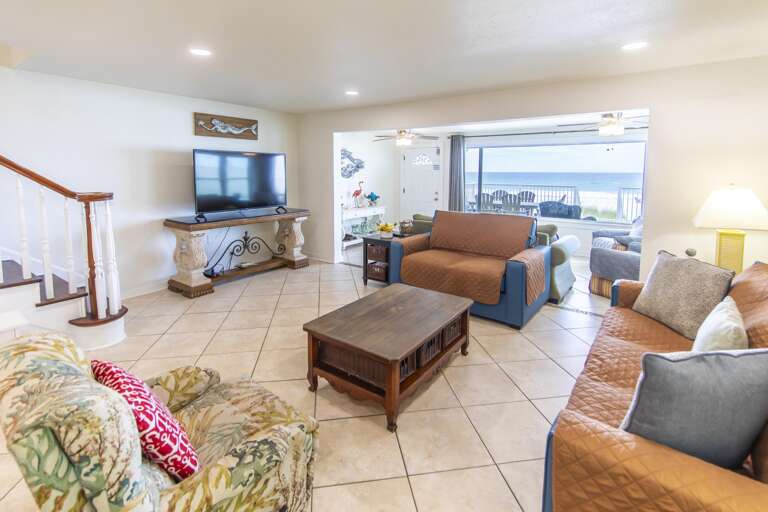 Tile floor living room with patterned armchair, leather-covered sofas, wooden coffee table, and ocean views beyond sliding glass.