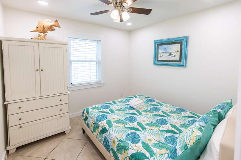 White wood armoire topped with a carved fish sculpture stands beside a window in the tile-floor bedroom.