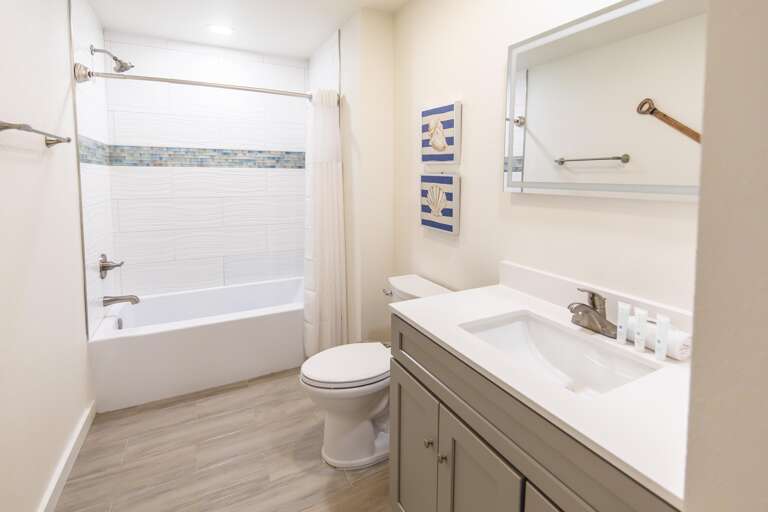 Full bathroom with wood-look tile flooring, white bathtub, brushed nickel fixtures, and nautical-themed wall art.
