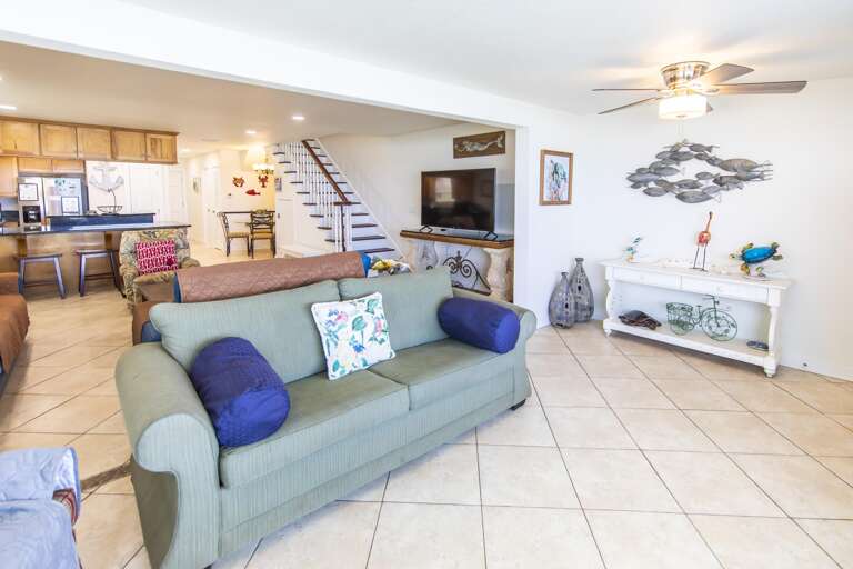 Living room with green sofa, blue pillows, tiled floors, coastal art, and a flat-screen TV on a carved wooden console.
