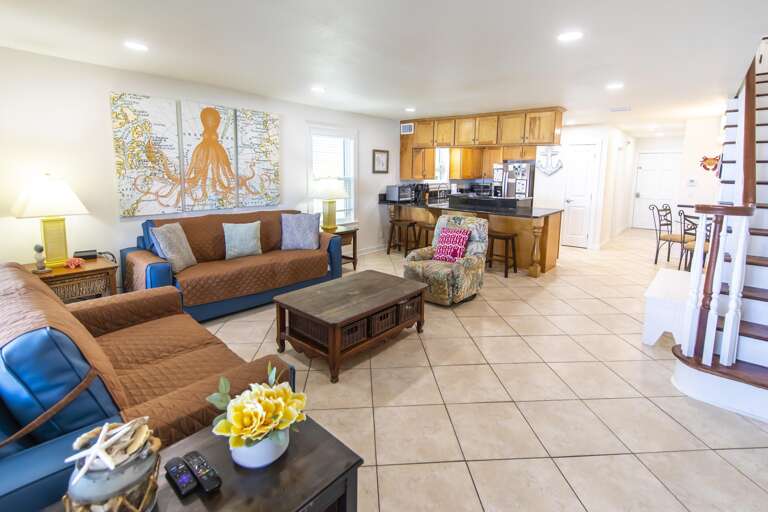 Open living area with tan tiled floors, wood coffee table, blue couches, and a kitchen bar with stools.
