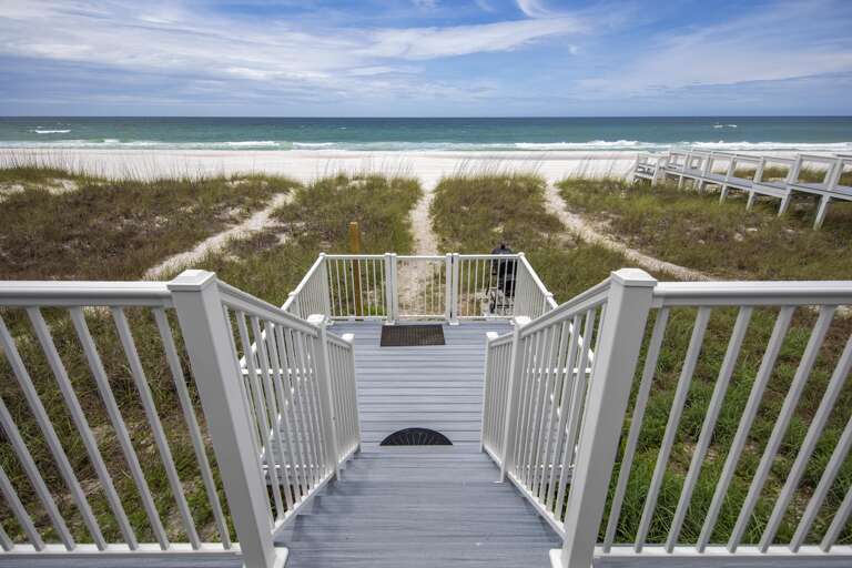 Gray composite stairs descend to sandy beach paths through dune grass under a partly cloudy sky.