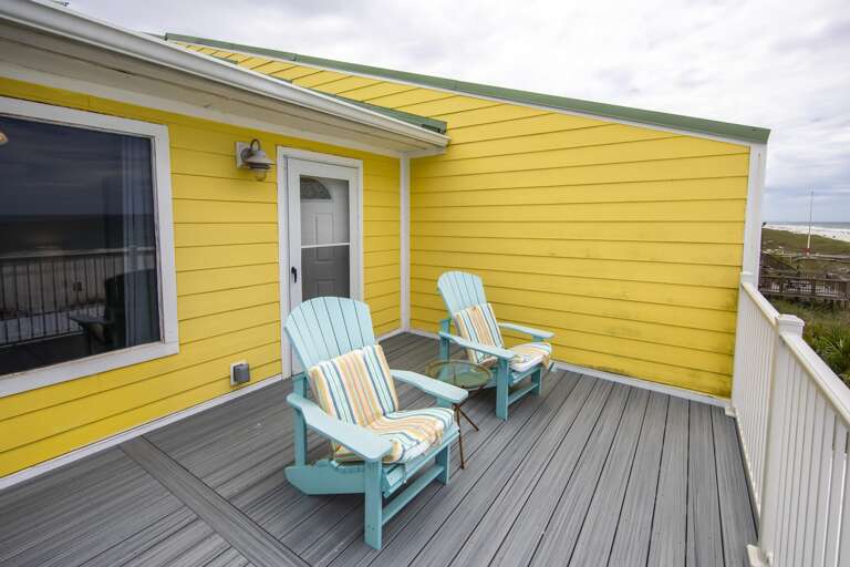 Corner balcony with gray composite flooring, yellow siding, two blue Adirondack chairs, and a small glass side table.