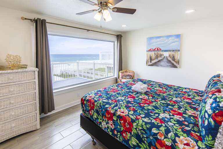 Large window in bedroom reveals ocean views above gray tile flooring and a white wicker dresser.