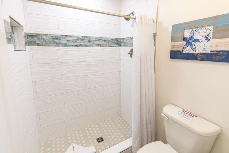 Walk-in shower with textured white tiles, blue mosaic accent strip, hex-patterned floor, and white shower curtain.