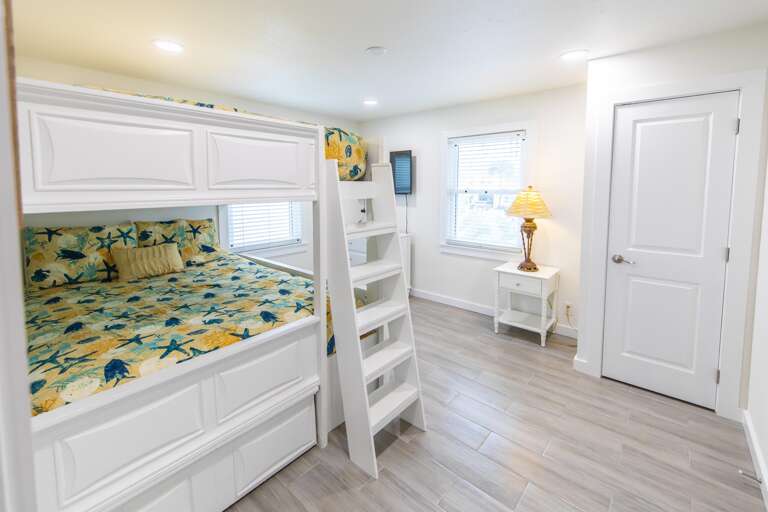 White bunk bed with a ladder stands against light gray wood-look tile flooring and white walls with two windows.