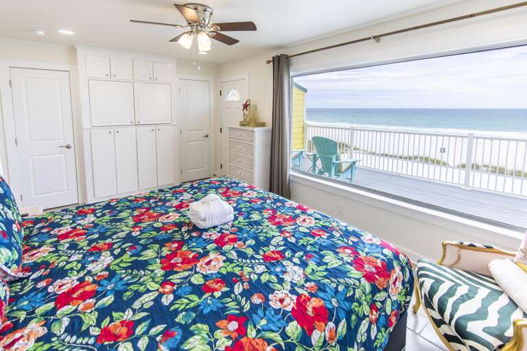 King bed with floral quilt faces a large window showing turquoise Adirondack chairs on the oceanfront deck.