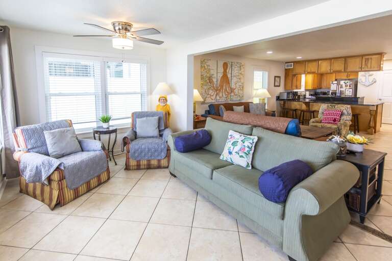 Tile flooring extends through the open living area, with soft green sofa, patterned armchairs, and coastal wall art.