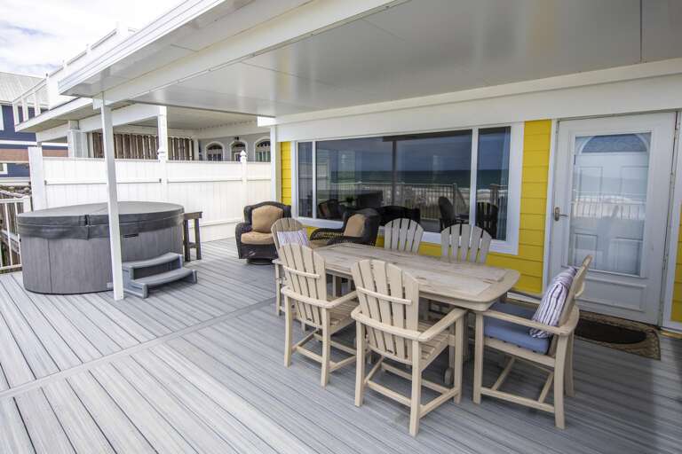 Outdoor dining area with light wood table and six chairs, next to a gray hot tub and ocean view window.