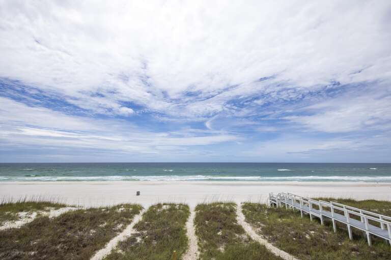 Sandy beach stretches beneath a partly cloudy sky, with a white wooden boardwalk crossing grassy dunes to the shore.