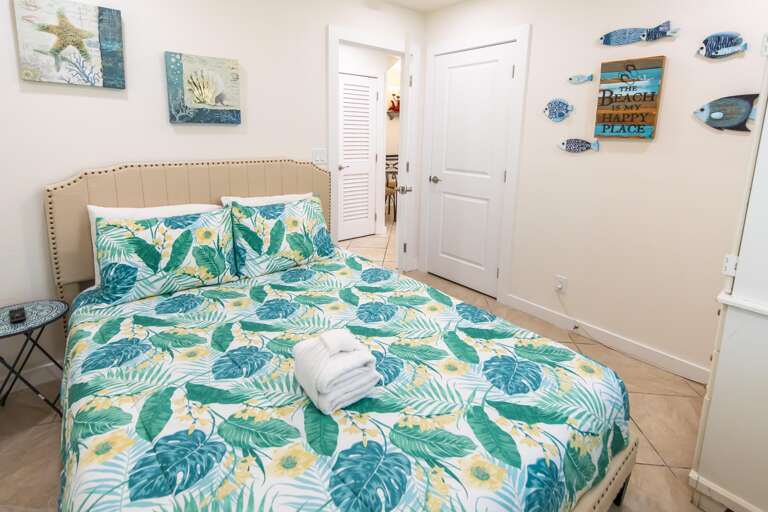 Bedroom with beige upholstered headboard, tropical leaf bedding, tile floor, and coastal-themed wall art.