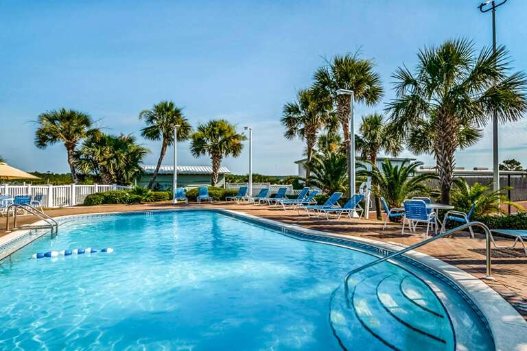 Sunlit pool with curved steps, surrounded by blue lounge chairs and palm trees under a clear sky.