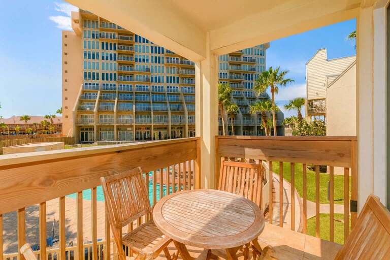 Balcony With Wooden Table, Chairs, Pool View, Palm Trees, And Buildings