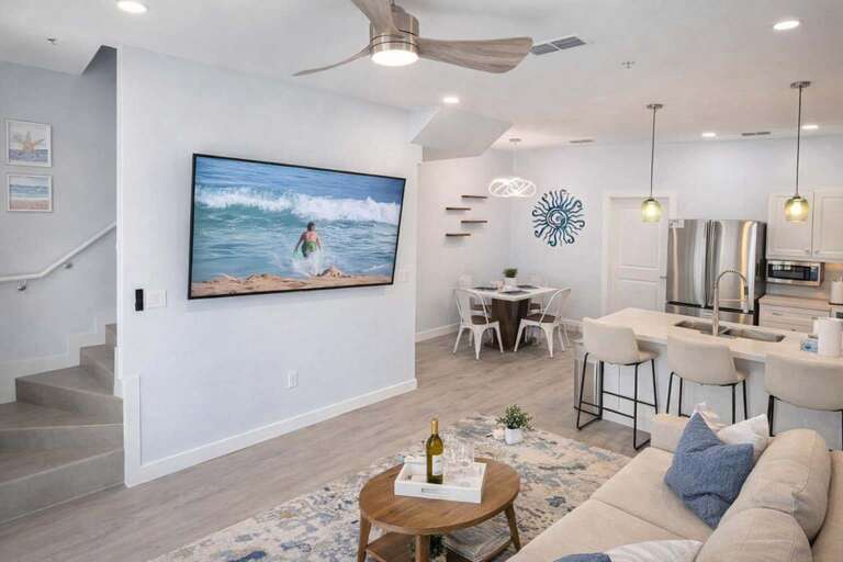 Open Living Area With Stairs, Wall Art, Large TV, Kitchen Island, And Dining Table