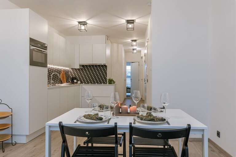 Compact Kitchen Leading To Light-drenched Dining Area With Black Chairs And Table Set For Two