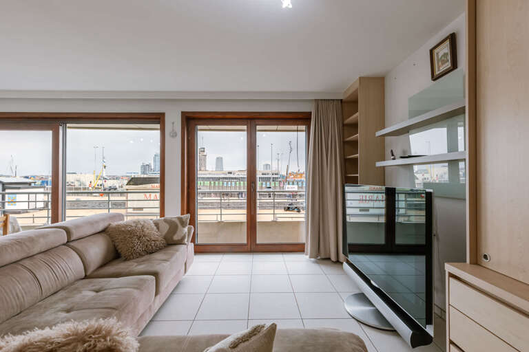 Living Room Leading To Balcony Overlooking Bustling Port, Showcasing Sofa, Shelves, And Sliding Doors