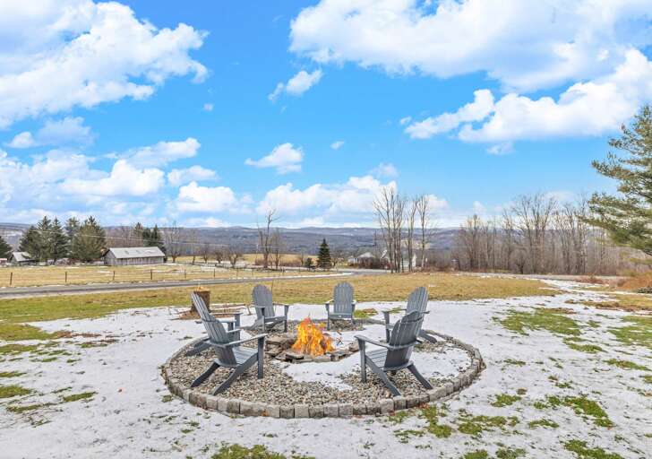 Chairs Circled Around A Fiery Furnace In A Frost-dusted Field Under A Fluffy Cloud-filled Sky