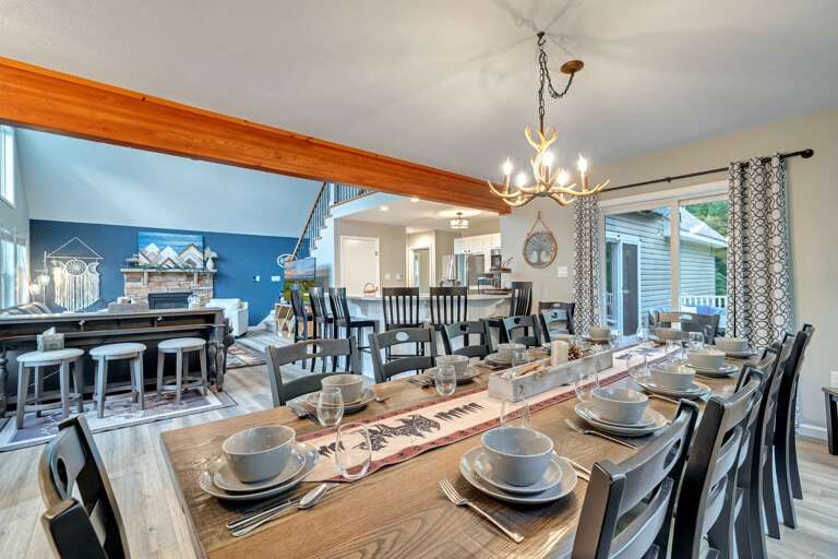 Dining Table With Place Settings, Chandelier Above, Open Kitchen, And Large Windows
