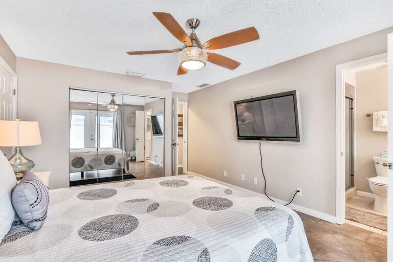 Relax in this bright beach-stay bedroom featuring a comfy bed, large TV, mirrored closets, and private ensuite access—perfect for unwinding after a day by the ocean.
