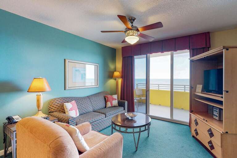 Bright beach-style living room with comfy seating, vibrant coastal décor, and sliding doors leading to a private balcony—perfect for relaxing with stunning ocean views.