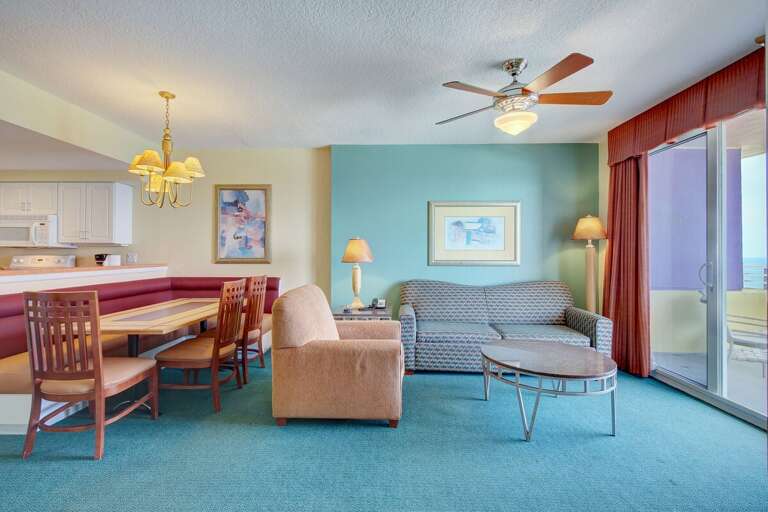 Colorful beach-style living and dining area with comfy seating, warm lighting, and sliding doors to a private balcony—perfect for unwinding after oceanfront adventures.