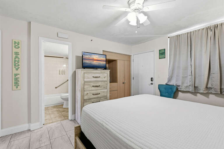 White-walled Room With Bed, Ceiling Fan, TV, And Adjoining Bathroom Door
