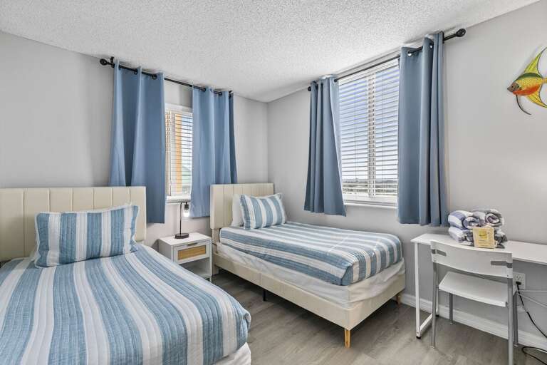 This bright and airy bedroom features two comfortable twin beds adorned with soft, coastal-themed bedding in blue and white stripes, perfectly complementing the room's seaside ambiance.
