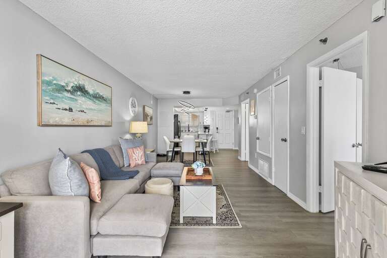 A plush sectional sofa with coral and ocean-blue accents provides comfort, while the large beach-themed artwork adds a serene, seaside touch.