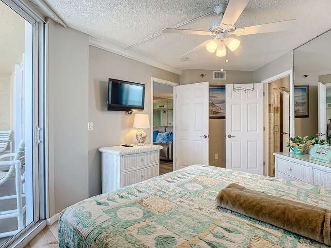 Bright bedroom offering a cozy bed, wall-mounted TV, mirrored accents, and direct balcony access for a perfect coastal escape.
