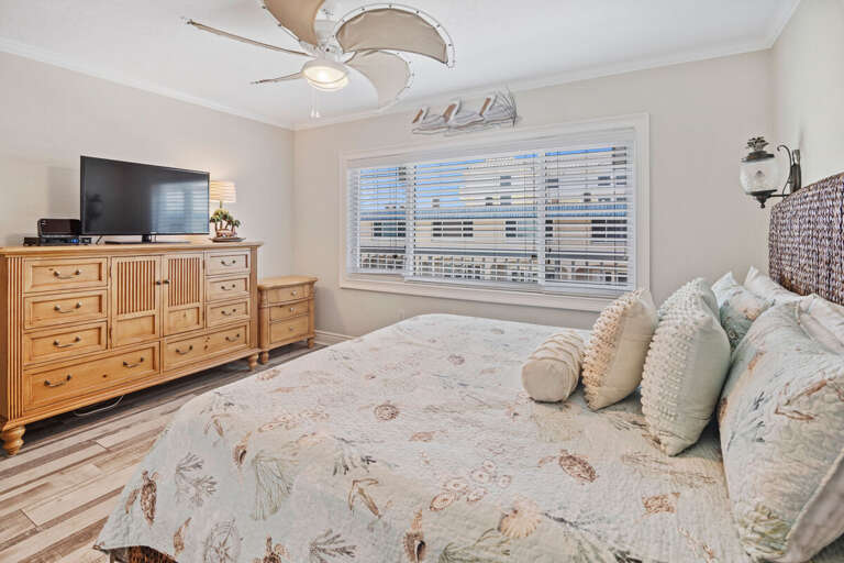 Rest easy after your beach stay in this inviting bedroom, featuring a plush bed with coastal bedding, natural wood furnishings, and a flat-screen TV for ultimate comfort and relaxation.