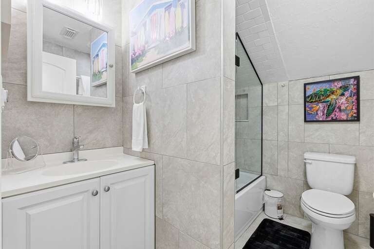 Clean coastal bathroom featuring a tub-shower combo, bright vanity, and beach-inspired artwork.