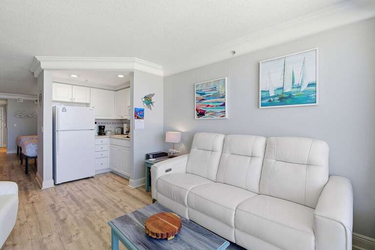 Relax in this comfortable beach stay living room featuring plush seating, coastal artwork, and an open layout leading to the kitchenette. A perfect spot to unwind after a fun-filled day by the beach.