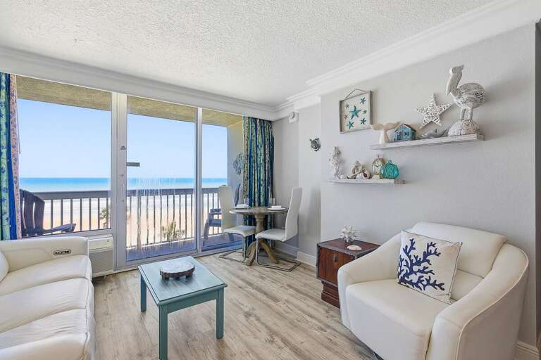 Stylish beach stay living area with cozy seating, coastal décor, and a dining nook by the window. Step onto the private balcony to take in beautiful oceanfront views just steps from the sand.