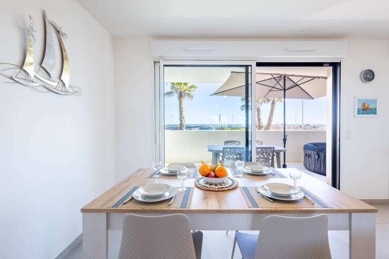 Dining Table With Plates And Fruit, Open Doors To Patio With Umbrella, Palm Trees, And Sea View