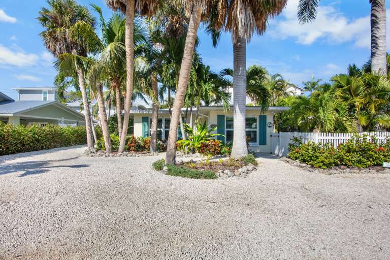 Tropical property entrance surrounded by swaying palm trees and lush landscaping with convenient gravel parking area.