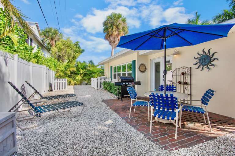 Your private outdoor oasis features a BBQ grill, comfortable seating under a bright blue umbrella, and lounge chairs on tropical landscaping - perfect for relaxing and dining outdoors.