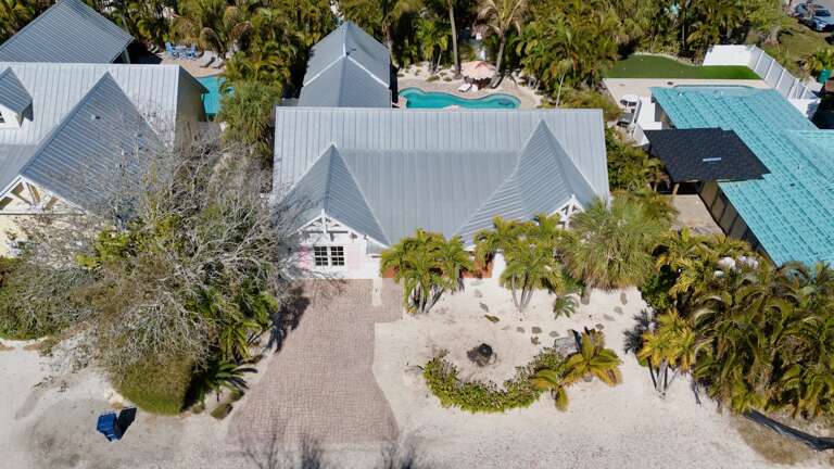 Aerial view of a tropical coastal property featuring charming architecture surrounded by swaying palm trees and neighboring pools in a serene island setting.