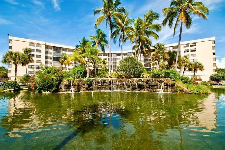 Tropical resort building with waterfront setting, featuring lush palm trees and landscaped grounds under bright blue skies.