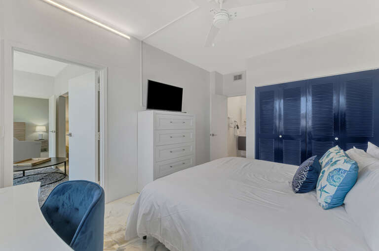 Drift off in your serene bedroom featuring crisp white linens, ocean-blue accents, and modern amenities including TV and ample storage.
