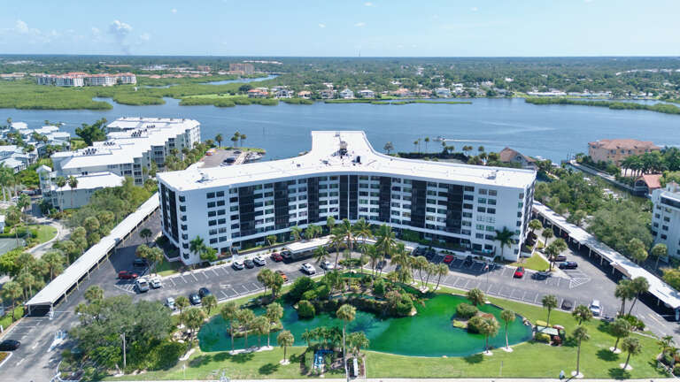 Tropical resort building with waterfront setting, featuring lush palm trees and landscaped grounds under bright blue skies.