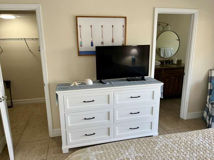 Your spacious bedroom features a white dresser with TV, coastal art, and convenient access to both walk-in closet and ensuite bathroom.