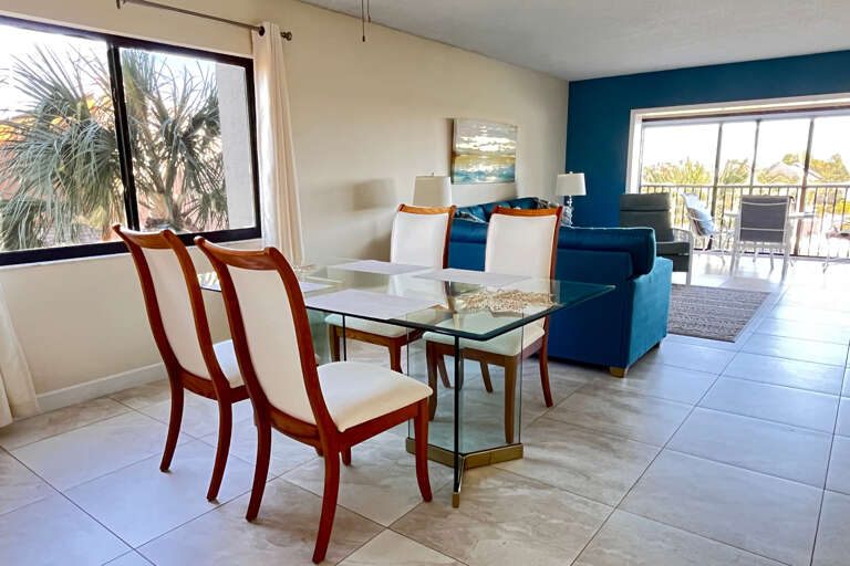 Unwind in your bright, coastal living space with modern glass dining and plush navy seating overlooking tropical palms.
