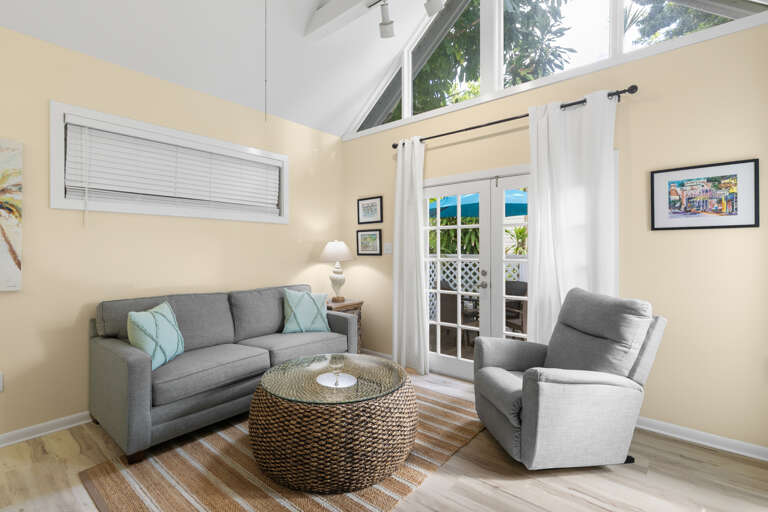 Relax in your sunlit living room where soft gray furnishings and soaring ceilings create an airy, welcoming retreat. Glass doors open to glimpses of tropical greenery beyond, inviting you to unwind in coastal comfort.