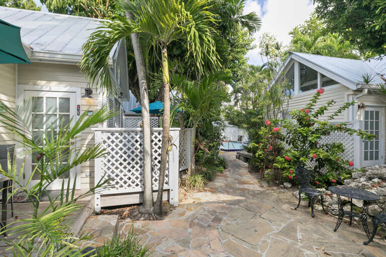 A charming cottage-style property nestled in lush tropical gardens with vibrant flowering plants and swaying palms. Stone pathways wind between private units, creating a tranquil island retreat.