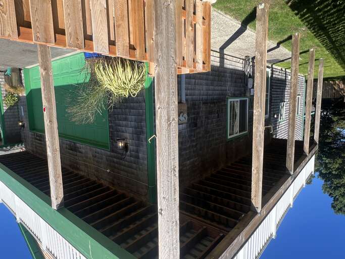 A unique floating rental property with dark wood siding and a bright green door, nestled on calm water. The structure sits on wooden pilings with a deck, surrounded by lush grass and trees along the shore.