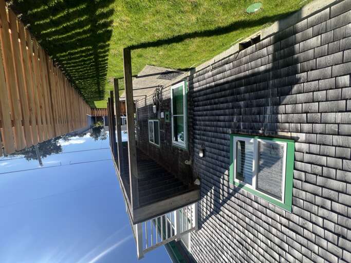 The property features dark shingle siding with bright green-trimmed windows and a private deck overlooking a lush green lawn enclosed by wooden fencing, creating a peaceful outdoor retreat.